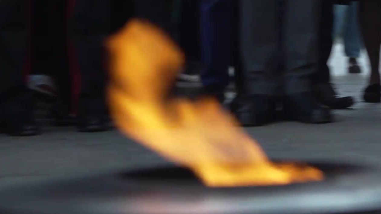 Gros plan de la flamme du Soldat inconnu ravivée, entourée de gerbes de fleurs blanches et bleues, sous l’Arc de Triomphe.