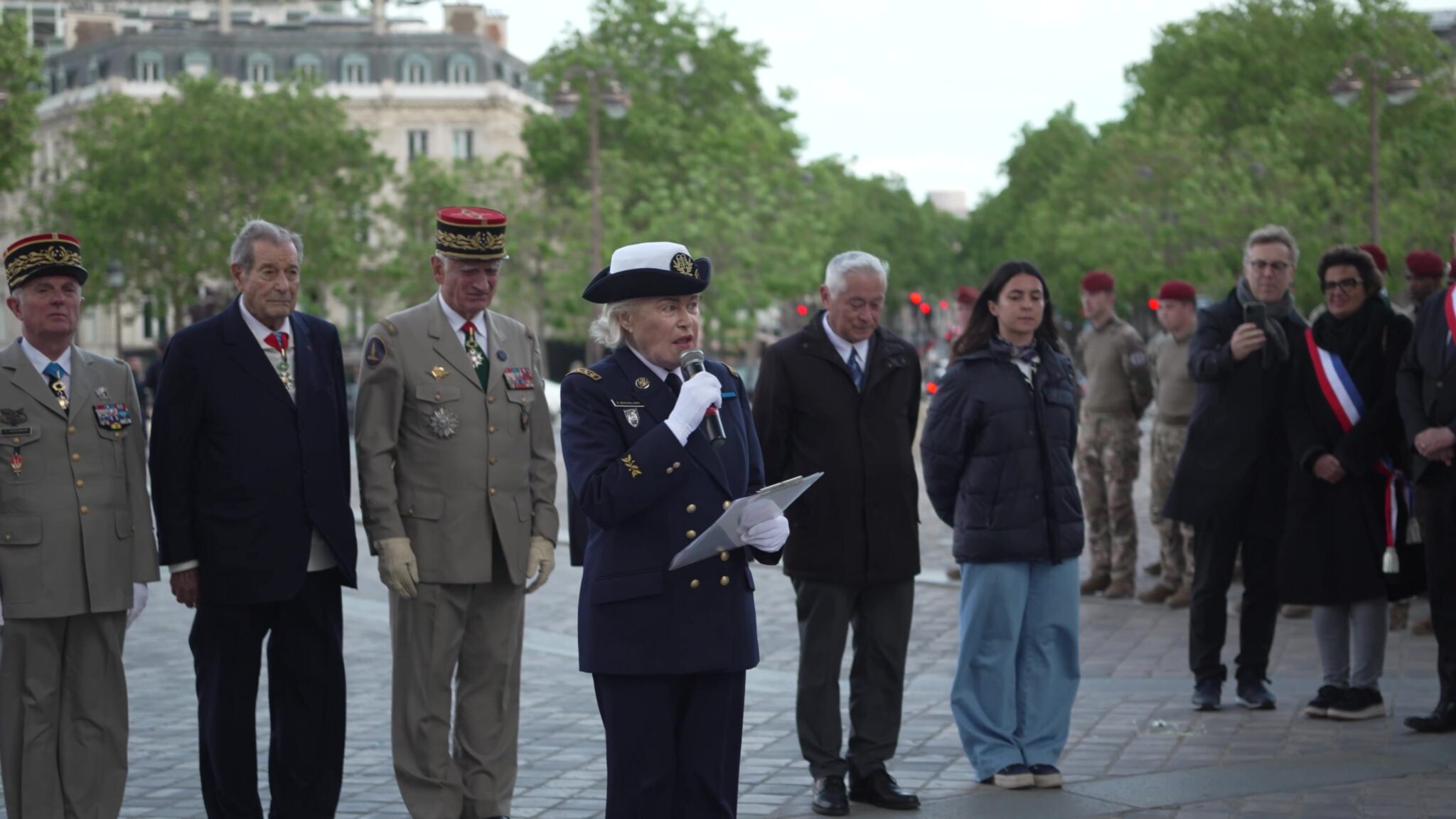 Une intervenante en uniforme prend la parole au microphone, entourée d’anciens combattants décorés et de militaires, sur l’avenue menant à l’Arc de Triomphe, lors de la cérémonie du 8 mai 2026.